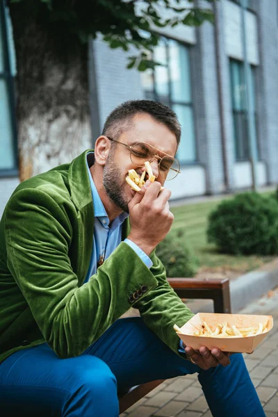 Portrait d'homme en veste de velours vert mangeant des frites assis sur un banc dans la rue — Photo de stock