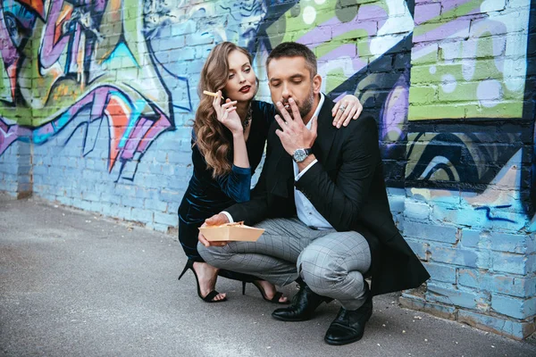 Couple en tenue à la mode avec des frites assis près du mur avec des graffitis sur la rue — Photo de stock