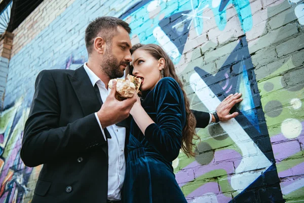 Low angle view of couple in luxury clothing with hot dogs standing near graffiti on wall on street — Stock Photo