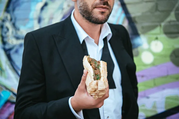 Vue partielle de l'homme en veste noire à la mode tenant hot dog dans la main sur la rue — Photo de stock