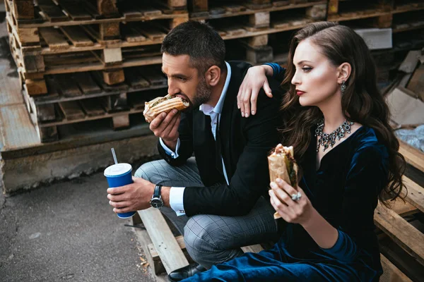 Vue latérale du couple en vêtements de luxe avec hot-dogs et soda assis sur une palette en bois dans la rue — Photo de stock