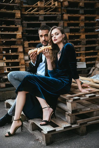 Vue latérale du couple en vêtements de luxe avec hot-dogs assis sur une palette en bois dans la rue — Photo de stock