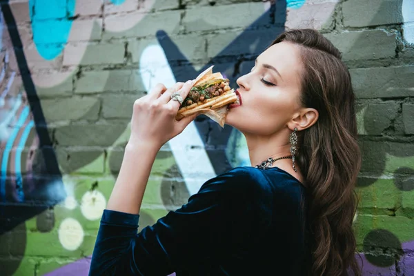 Side view of beautiful woman in stylish clothing eating hot dog on street — Stock Photo