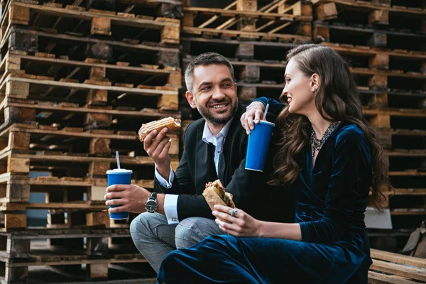 Вид сбоку улыбающейся пары в роскошной одежде с хот-догами и газировкой, сидящей на деревянном поддоне на улице — стоковое фото