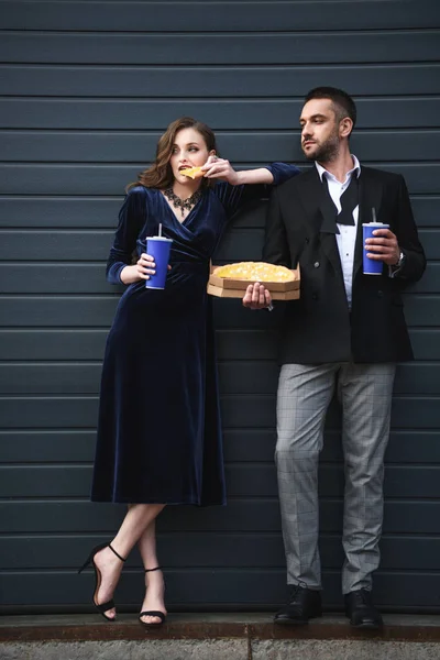 Couple en tenue de luxe avec boissons gazeuses et pizza italienne posant dans la rue — Photo de stock
