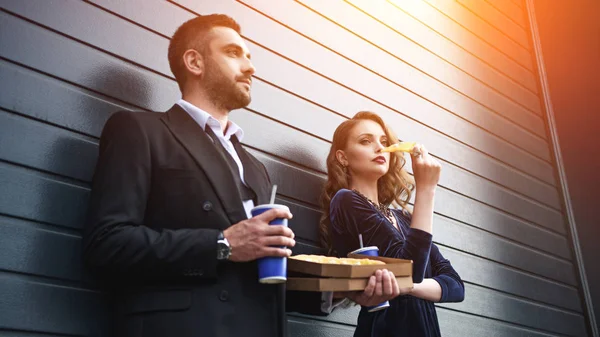 Vista lateral de pareja en traje de lujo con bebidas gaseosas y pizza italiana en la calle - foto de stock