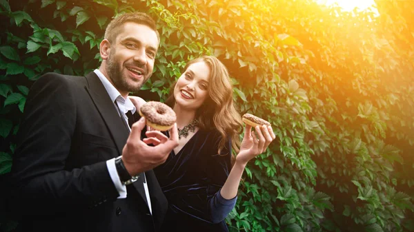 Portrait of smiling fashionable couple with chocolate doughnuts with green foliage behind — Stock Photo