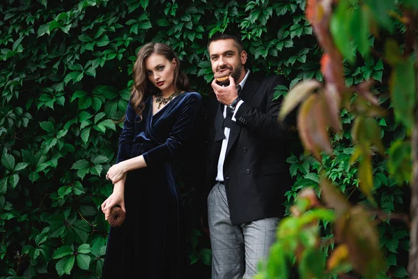 Pareja de moda con donas de chocolate con follaje verde detrás mirando a la cámara - foto de stock