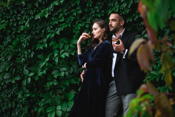 Couple à la mode avec beignets au chocolat avec feuillage vert derrière regarder loin — Photo de stock