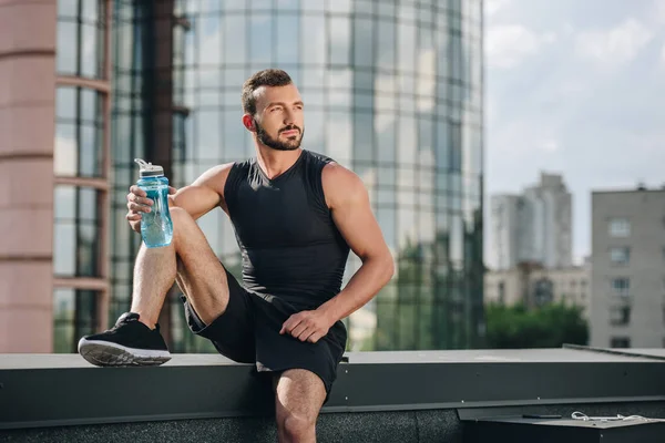 Красивый спортсмен отдыхает и держит спортивную бутылку воды на крыше — стоковое фото