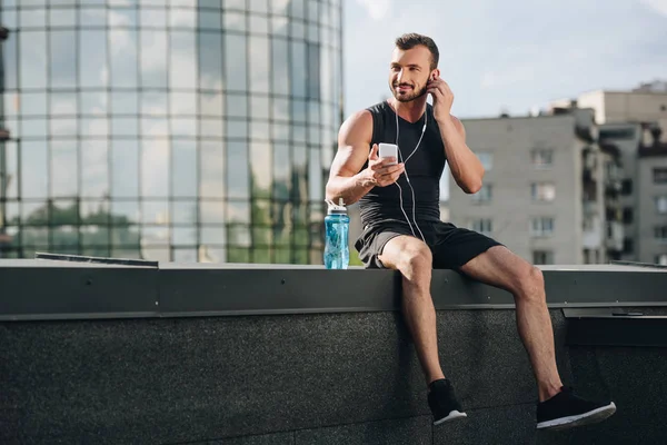 Handsome cheerful sportsman listening music and using smartphone on roof — Stock Photo