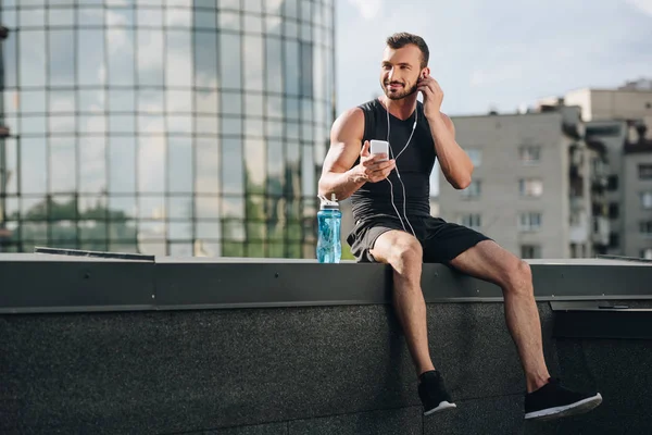 Beau sportif joyeux écoutant de la musique et utilisant un smartphone sur le toit — Photo de stock