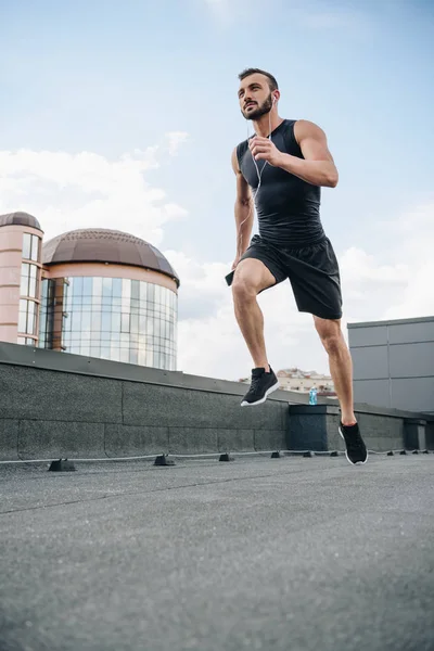 Vista de ángulo bajo del deportista guapo corriendo y escuchando música con teléfono inteligente en el techo - foto de stock