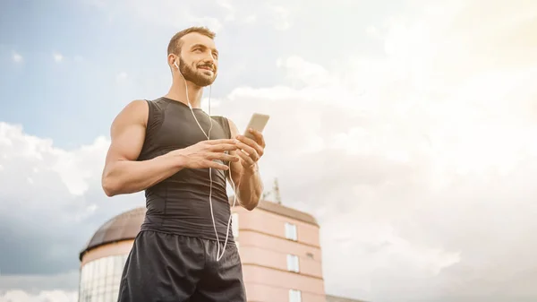 Vista de ángulo bajo de deportista guapo escuchando música con teléfono inteligente y mirando hacia otro lado en el techo - foto de stock