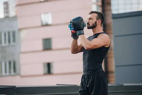 Seitenansicht eines gutaussehenden Boxers in Handschuhen, der Wasser aus einer Sportflasche auf dem Dach trinkt — Stockfoto