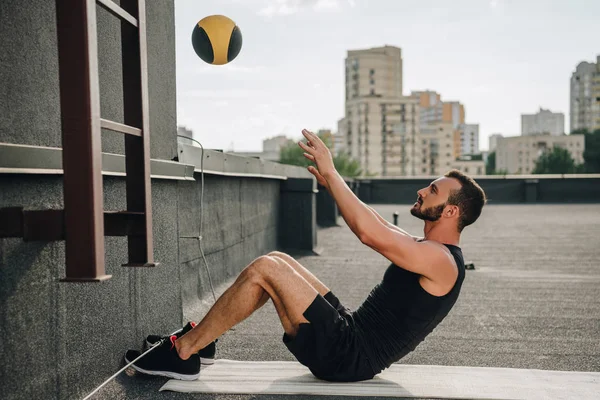Vue latérale d'un beau sportif faisant des redressements assis avec une boule de médecine sur un tapis de yoga sur le toit — Photo de stock