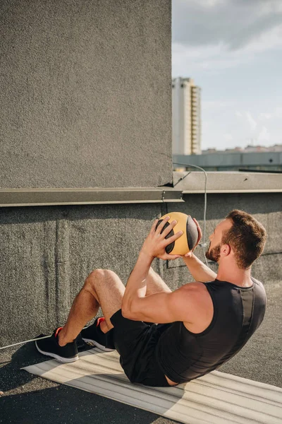Deportista haciendo sit ups con bola de medicina en yoga mat en techo - foto de stock