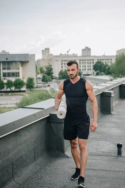 Handsome sportsman walking with yoga mat on roof — Stock Photo