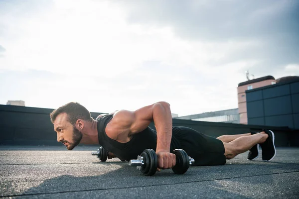 Side view of handsome sportsman doing push ups with dumbbells on roof — Stock Photo