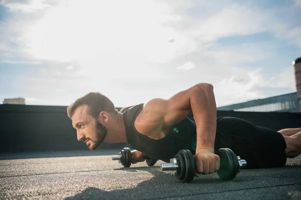 Side view of handsome sportsman doing plank on dumbbells on roof — Stock Photo