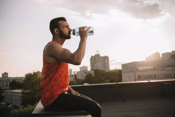 Vista lateral del deportista guapo beber agua de la botella de deporte en el techo — Stock Photo