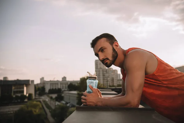 Seitenansicht eines gut aussehenden Sportlers, der eine Flasche Wasser in der Hand hält und sich an ein Geländer auf dem Dach lehnt — Stockfoto