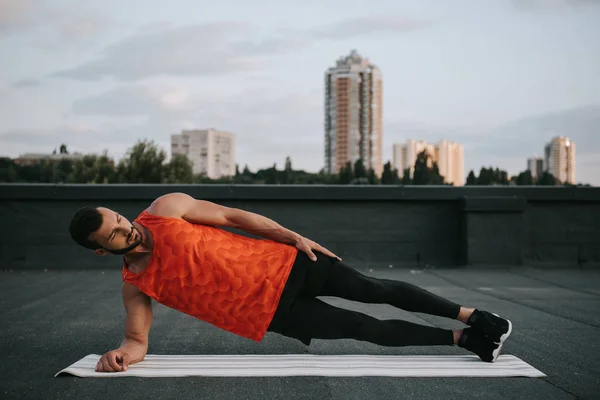 Handsome sportsman doing side plank on yoga mat on roof — Stock Photo