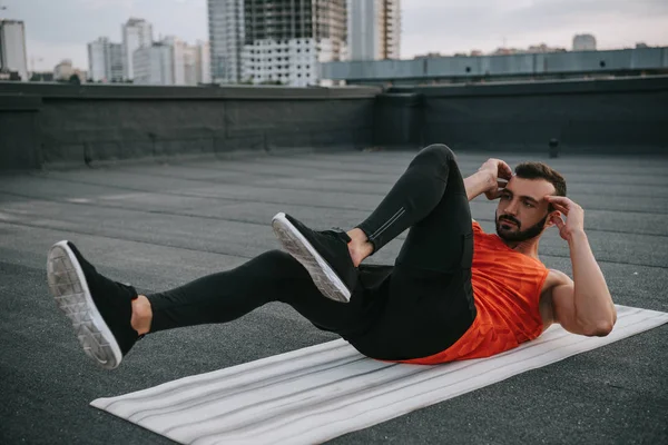 Handsome sportsman doing sit ups on yoga mat on roof — Stock Photo