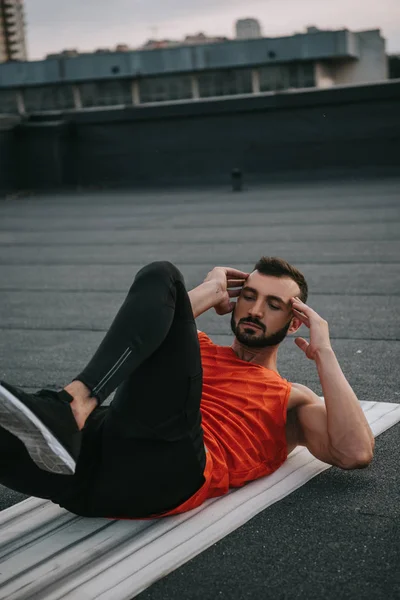 Handsome athletic sportsman doing sit ups on yoga mat on roof — Stock Photo