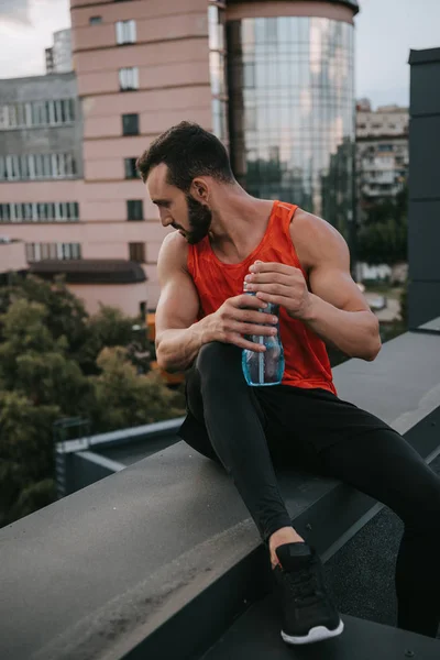 Deportista guapo sentado en la barandilla del techo con botella de agua y mirando hacia abajo - foto de stock