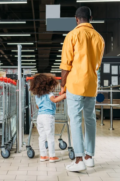 Vista trasera del hombre afroamericano de pie con su hija cerca del carrito de compras en el hipermercado - foto de stock