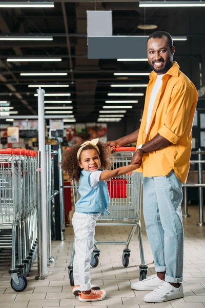 African american man holding hands of daughter near shopping trolley in grocery store — Stock Photo