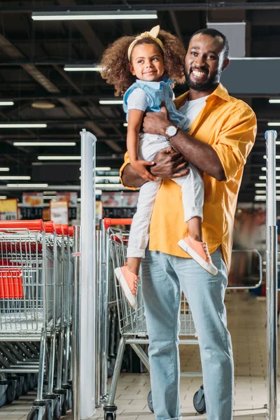 Smiling african american man embracing and holding little daughter in supermarket — Stock Photo