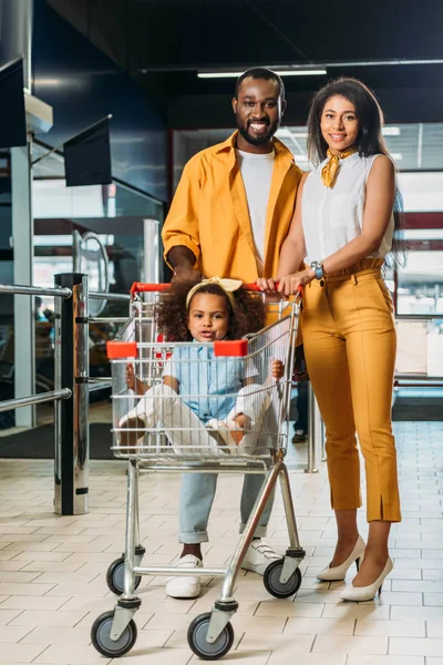 Adorable african american kid sitting in shopping trolley while her parents standing near in supermarket — Stock Photo