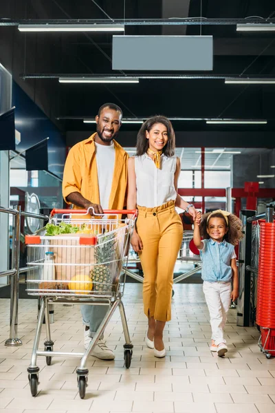 Sonriente afroamericano hombre llevando carrito de compras mientras su esposa toma de la mano de su hija en el supermercado - foto de stock
