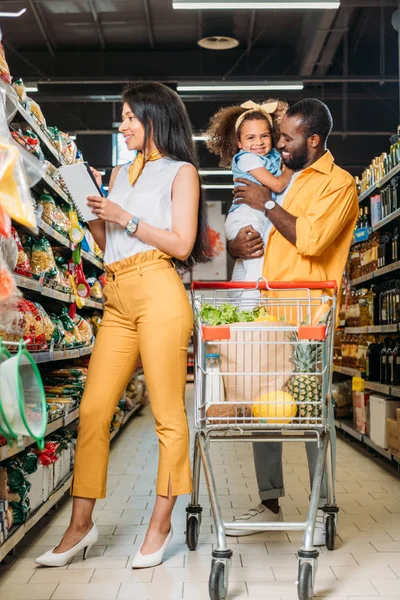 Sorridente uomo africano americano che tiene la figlia mentre sua moglie sceglie il cibo con la lista della spesa nel supermercato — Foto stock