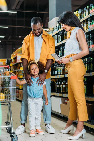 Feliz africano americano pareja con hija de pie cerca de carrito de compras en supermercado - foto de stock