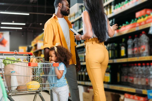 Foco seletivo do casal afro-americano escolhendo comida enquanto sua filha está com carrinho de compras no hipermercado — Fotografia de Stock