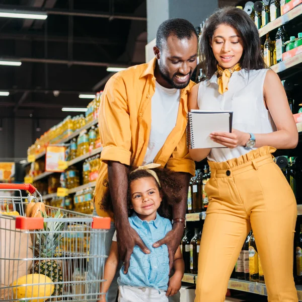 Happy african american family with daughter looking at shopping list in supermarket — Stock Photo