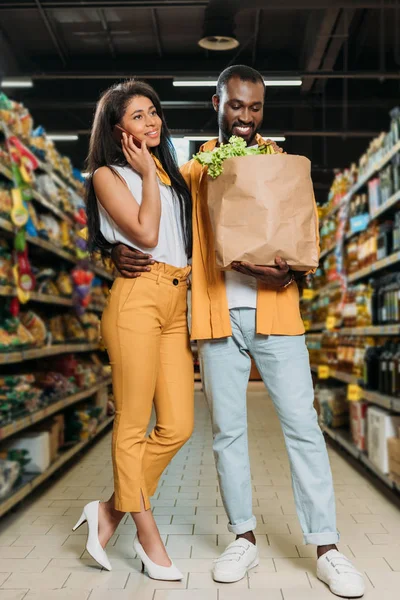 Sonriente afroamericano hombre sosteniendo bolsa de papel con comida y abrazando novia mientras ella habla en smartphone - foto de stock