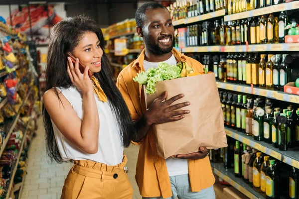 Sonriente afroamericano hombre sosteniendo bolsa de papel con comida mientras su novia habla en el teléfono inteligente - foto de stock
