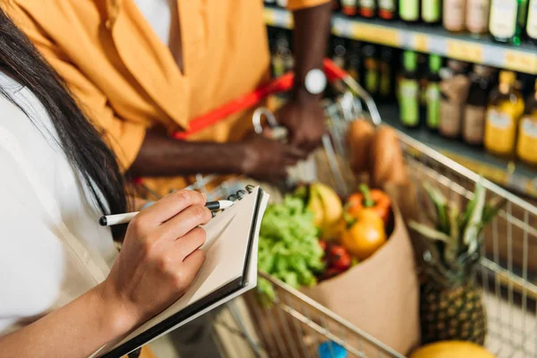 Immagine ritagliata di donna marcatura lista della spesa mentre il suo ragazzo in piedi vicino con carrello della spesa nel supermercato — Foto stock