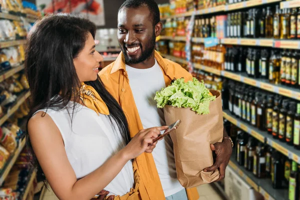 Junges afrikanisch-amerikanisches Paar mit Smartphone und Papiertüte mit Lebensmitteln im Supermarkt — Stockfoto