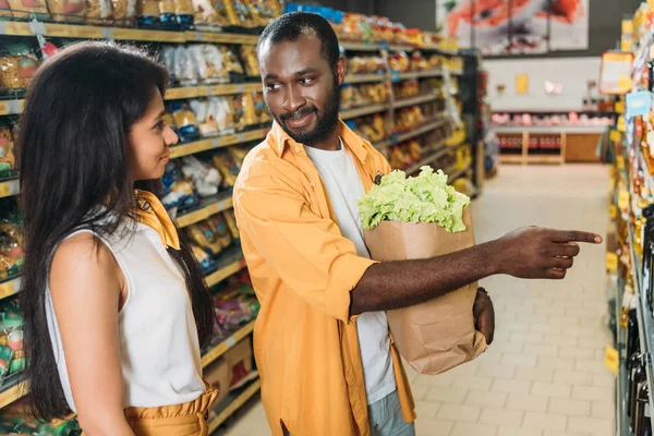 Uomo africano americano con sacchetto di carta con il cibo che punta alla fidanzata nel supermercato — Foto stock