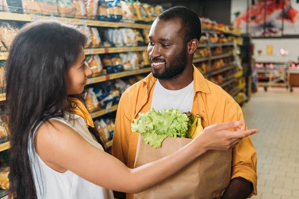Young african american woman pointing to boyfriend with paper bag with food in supermarket — Stock Photo