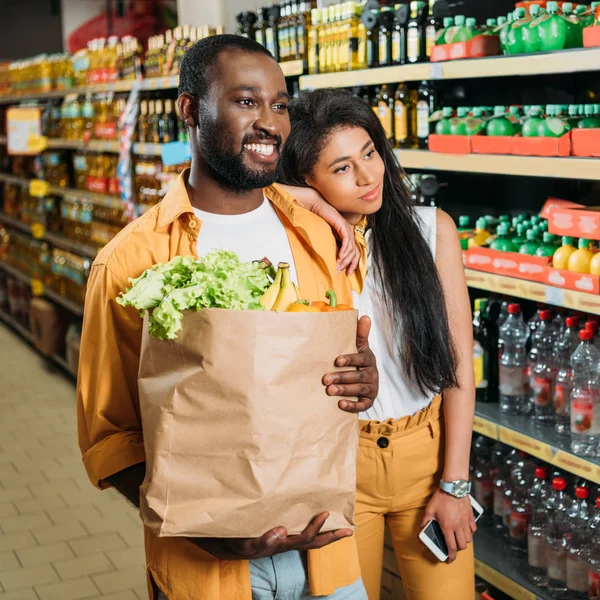 Усміхнена афро-американська пара з паперовим пакетом та смартфоном у продуктовому магазині — стокове фото