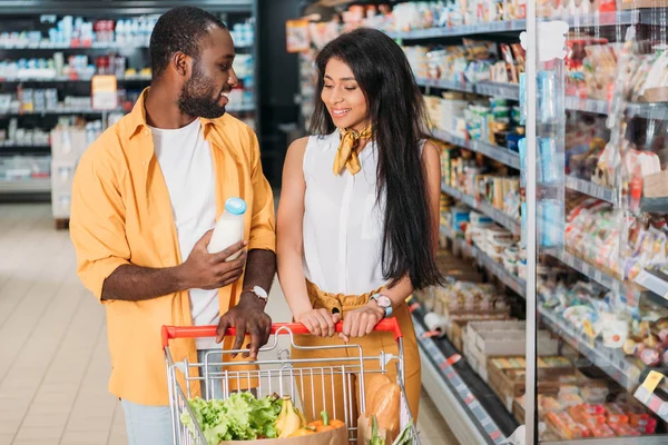 Joven afroamericano hombre mostrando leche a novia con carrito de compras en supermercado - foto de stock