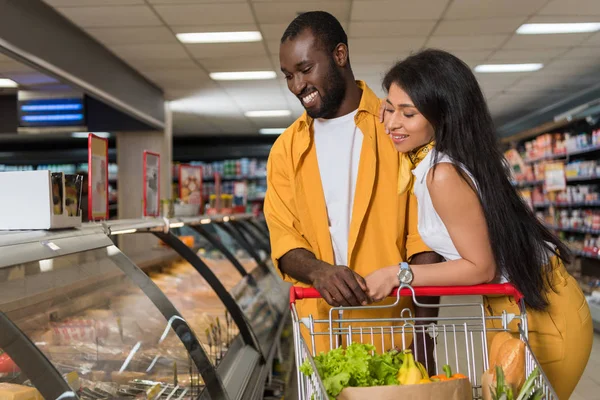 Sonriente pareja afroamericana con carrito de compras eligiendo comida en el supermercado - foto de stock