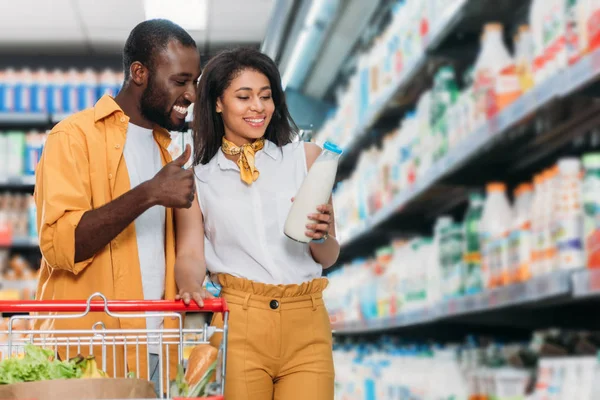 Sonriente afroamericano hombre haciendo el gesto de pulgar hacia arriba a la novia sosteniendo leche en el supermercado - foto de stock