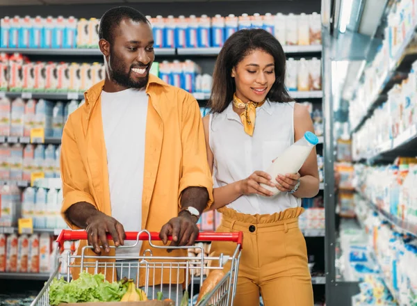 Sorridente coppia afro-americana con carrello della spesa che sceglie il latte al supermercato — Foto stock